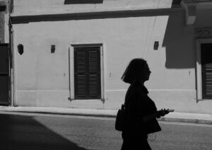 On A Mission shows a photo image in 35mm digital black and white of a woman in silhouette walking quickly on a sunny morning with a mobile phone on a Malta street.