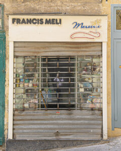 Vanishing Malta 2025 shows a 35mm digital colour image of a disused and delapitated shop with lots of items left behind in the display windows